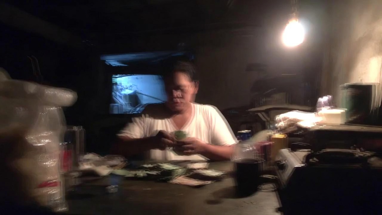 Filipino woman counting small folded bills at a cluttered kitchen table, dim warm light, strongly blurred.